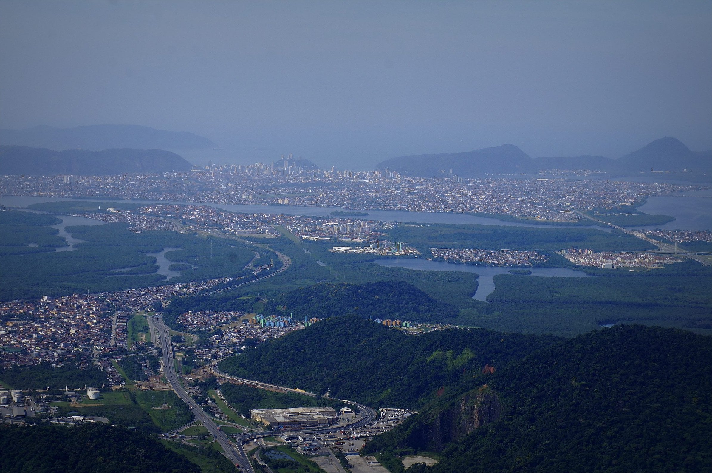 Vista do alto da Serra do Mar sobre Cubatão, com a Via Anchieta, os bairros à beira dos rios e manguezais e o estuário de Santos ao fundo