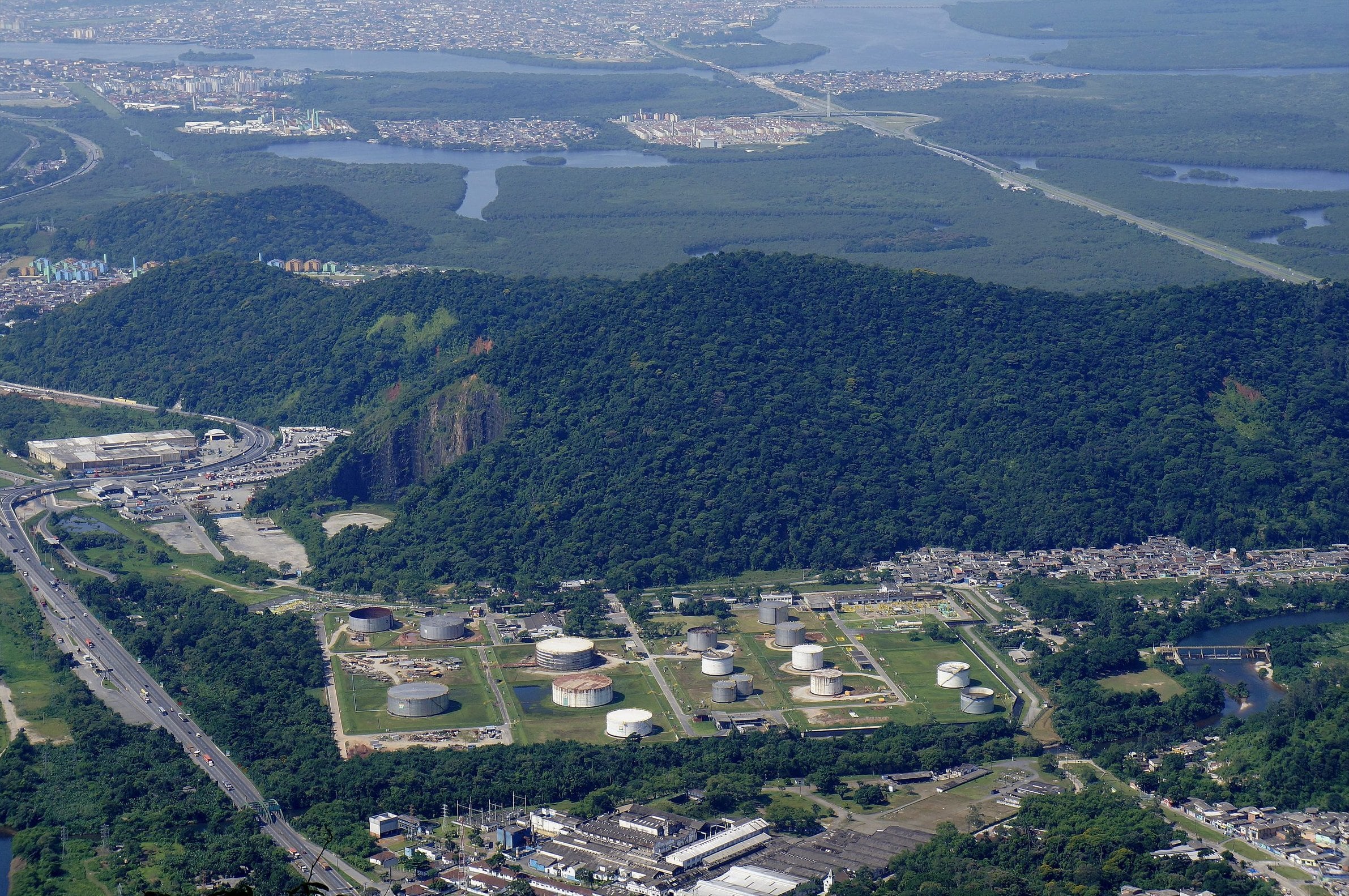 Vista ensolarada de Cubatão a partir da Serra do Mar, com morros verdes, a área industrial à beira da Via Anchieta e os manguezais ao fundo