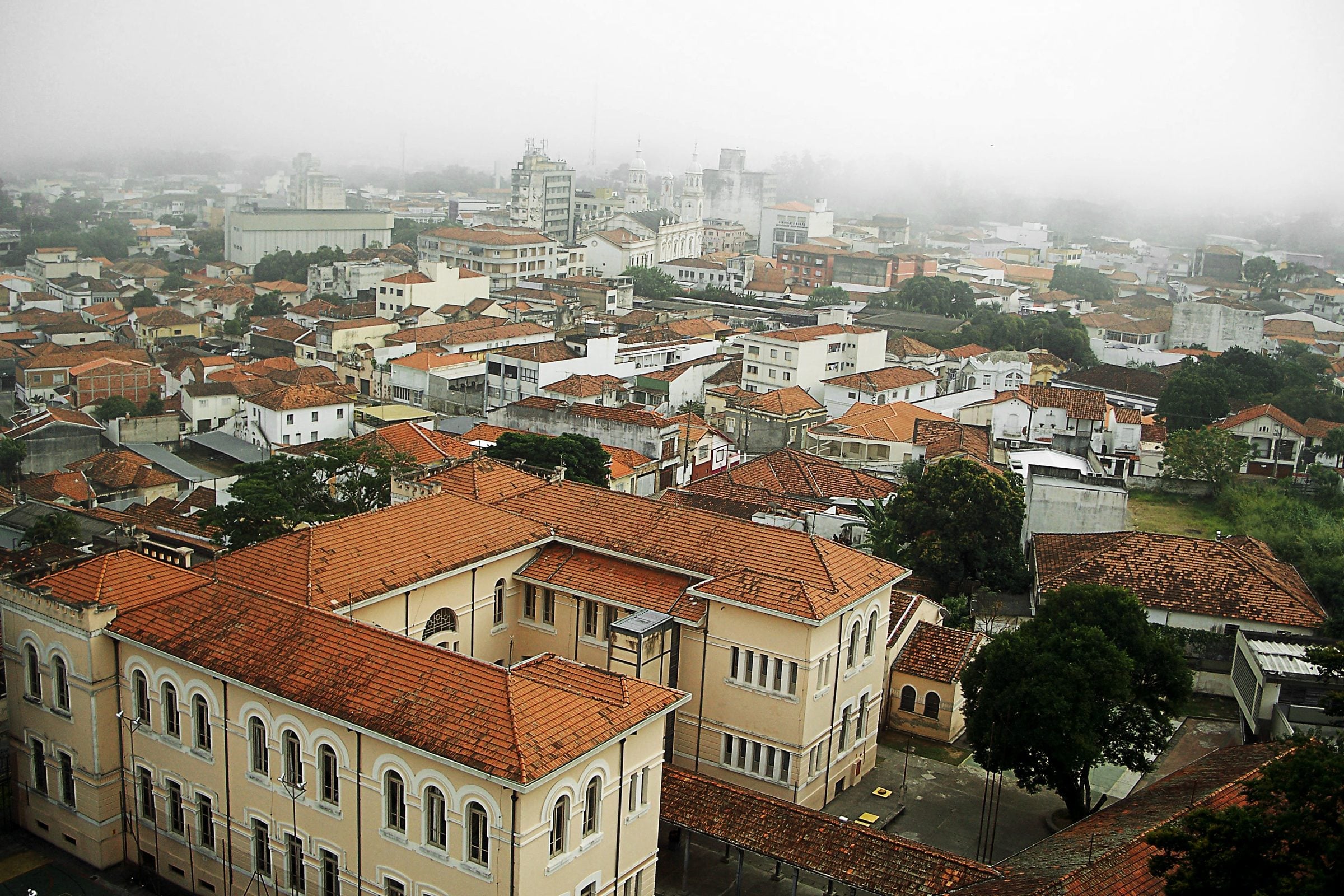 Vista do alto do centro de Guaratinguetá numa manhã de neblina, com os telhados e o prédio histórico da Escola Estadual Doutor Flamínio Lessa em primeiro plano