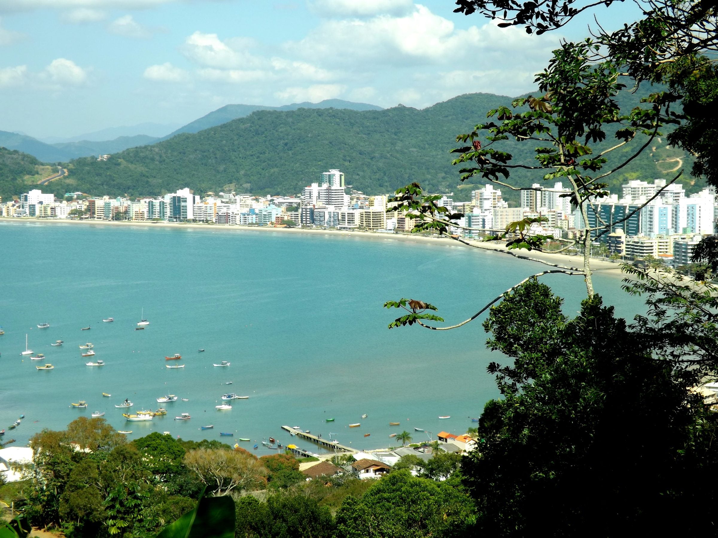 Vista do mirante sobre a enseada de Itapema, com barcos na água azul, os prédios da Meia Praia e os morros verdes ao fundo
