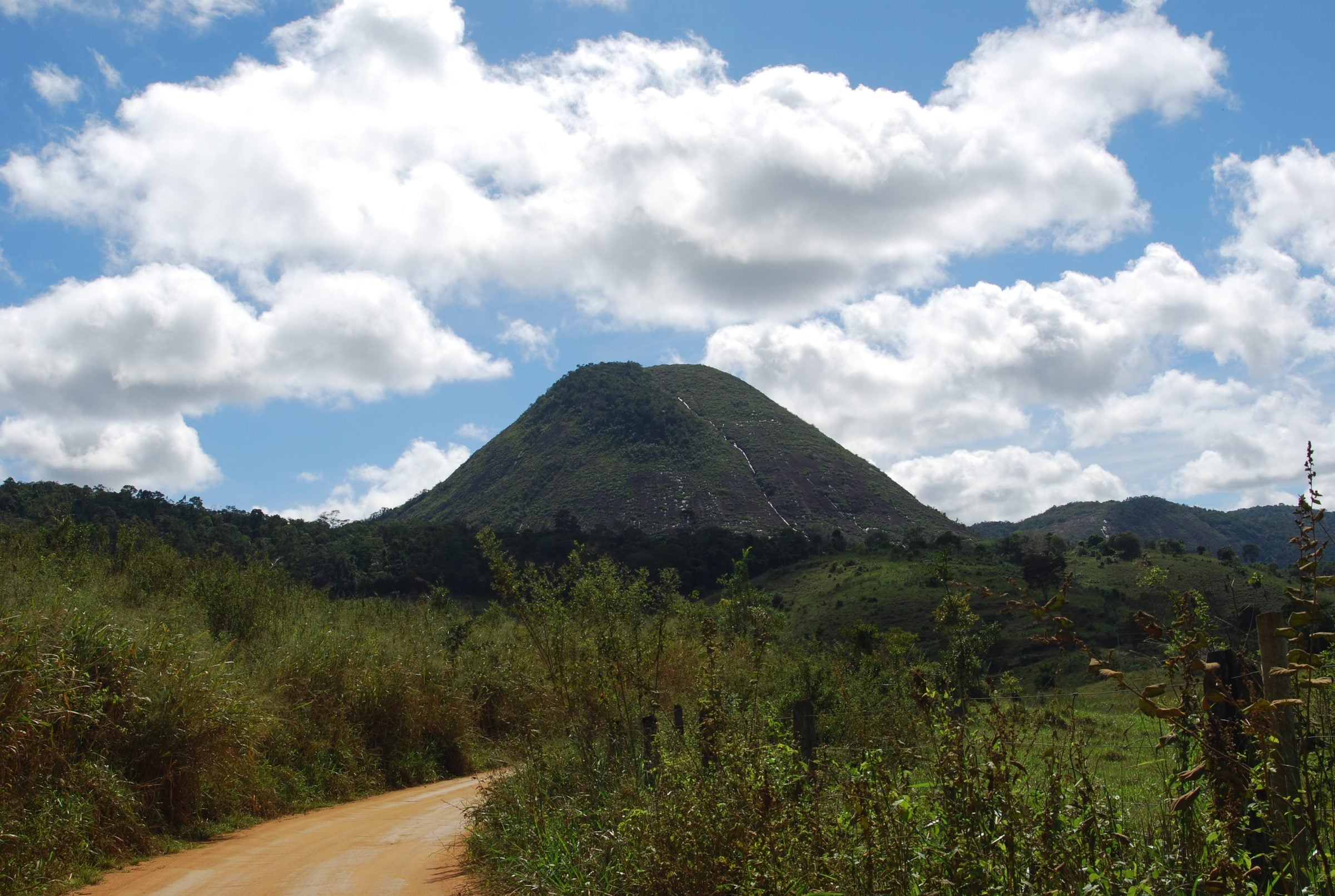 Paisagem rural de Novo Cruzeiro, com estrada de terra, mata e um morro de pedra sob céu azul